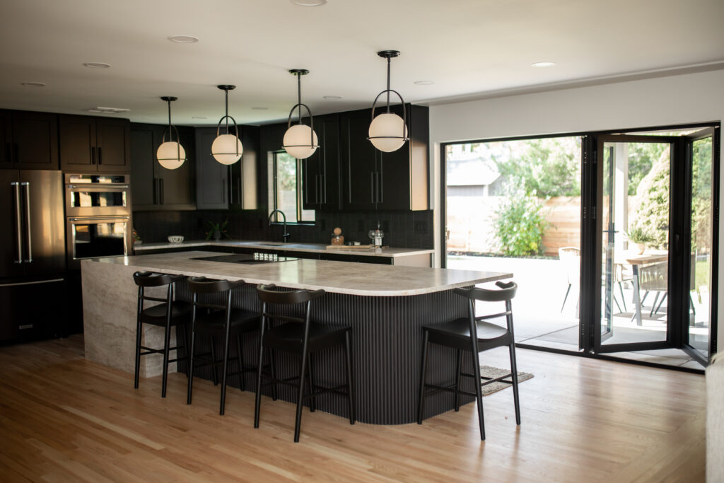 Modern kitchen renovation with black cabinets, white countertops, a central island with bar stools, and pendant lighting, seamlessly connecting to an outdoor space through large sliding doors.