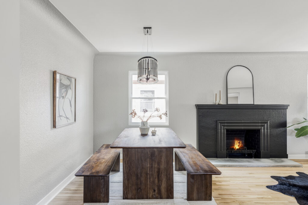 A minimalist dining room with a wooden table and two benches, illuminated by a contemporary hanging light fixture. A fireplace with a black mantle and a mirror above it is set in a light gray textured wall. A framed artwork hangs on the adjacent wall. Light wood flooring, window in background.