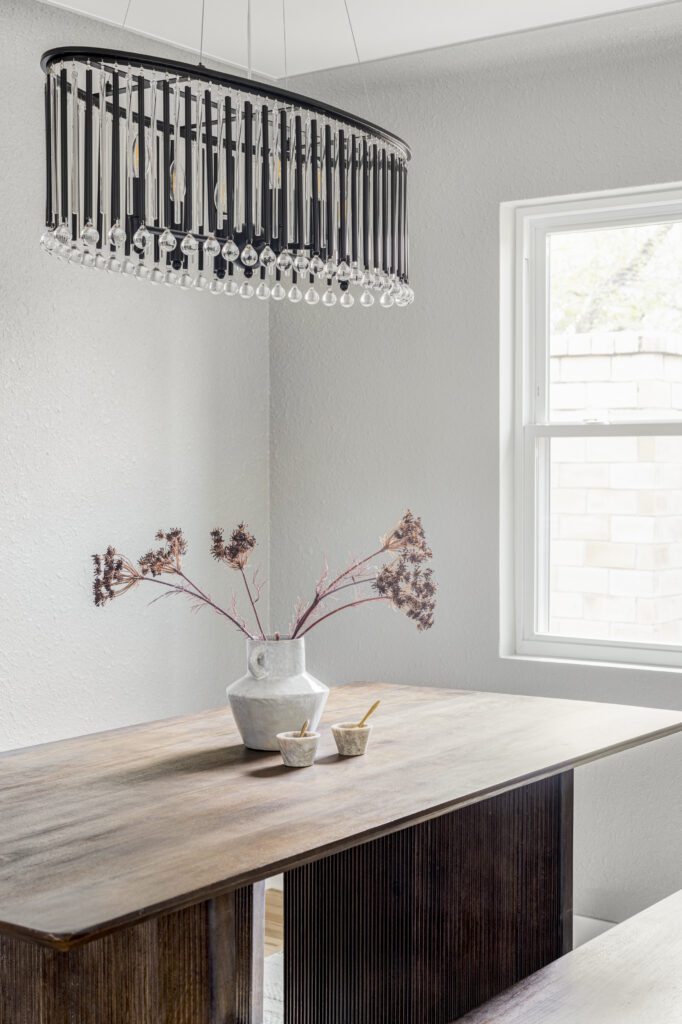 A minimalist dining area features a sleek wooden table with a white ceramic vase holding dried flowers. Two small bowls with wooden spoons sit on the table. A modern chandelier with hanging crystals illuminates the space, which is bathed in natural light from a nearby window.