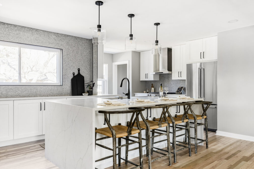 A modern kitchen with a large white marble island, four wooden bar stools, white cabinets, stainless steel appliances, and hanging pendant lights. A window on the left wall lets in natural light, and a stylish backsplash adorns the wall behind the sink area.