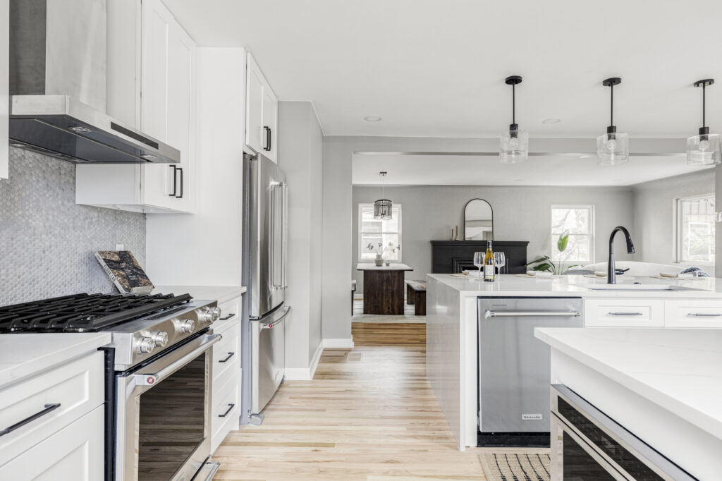 A modern kitchen with white cabinetry, stainless steel appliances, and a large central island. Hanging pendant lights provide illumination. The space opens up into a living area featuring a dark fireplace, light gray walls, and wood flooring throughout.