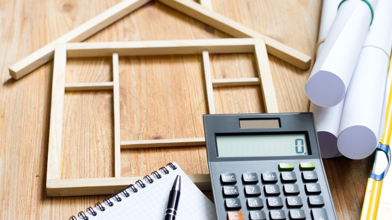 An arrangement on a wooden desk features a wooden frame shaped like a house, a spiral notebook with a pen, a calculator displaying zero, and rolled-up architectural plans. A tape measure is also partially visible. All tools for a home remodel planning process