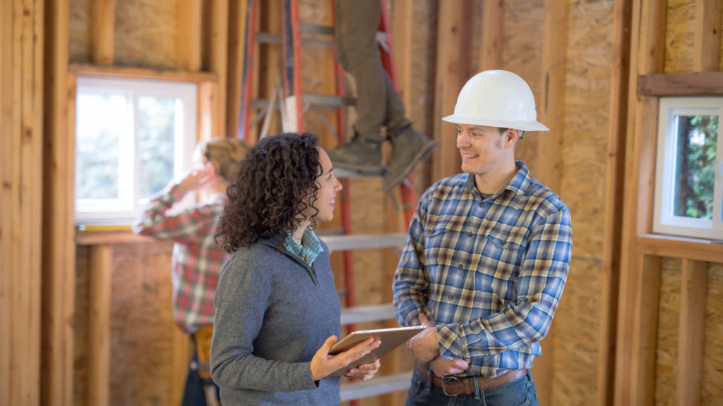 project manager or general contractor wearing a hardhat on a construction site of a home remodel. The project manager is speaking to the homeowner about the remodel process, while subcontractors work on ladders in background