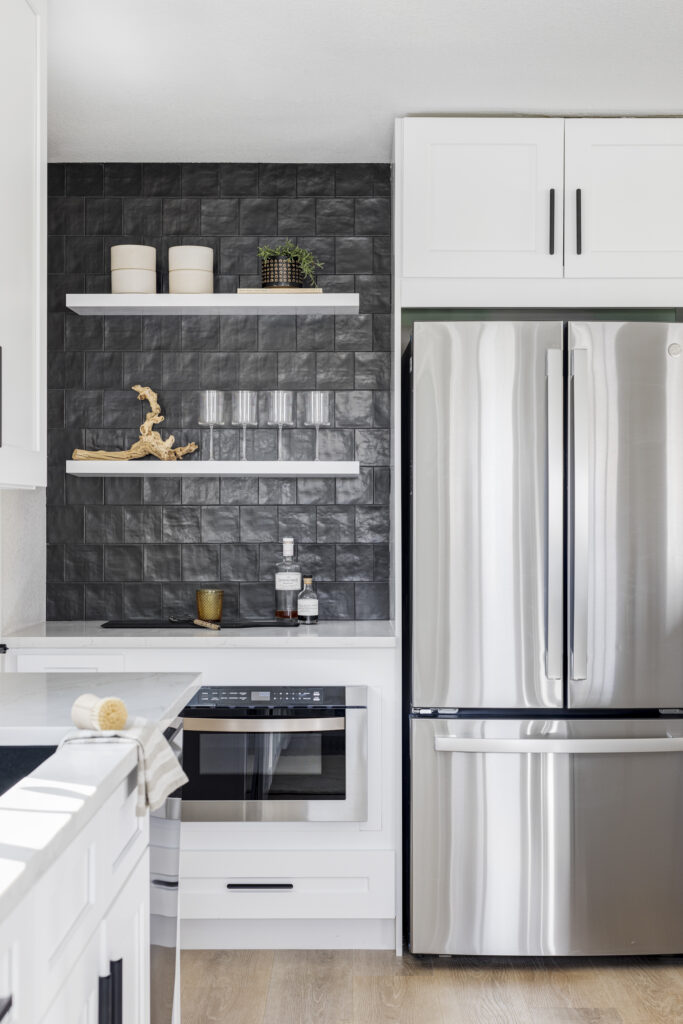 A modern kitchen with white cabinetry, stainless steel refrigerator, and a built-in oven below the countertop. The backsplash features black textured tiles with two floating shelves holding minimal decor, including jars, bottles, and plants. Light wood flooring.