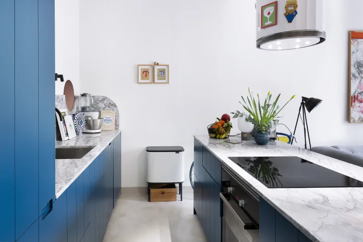 a modern kitchen with bright blue cabinets, marble countertop, and an integrated cooktop stove that is seamlessly part of the kitchen island. There are other modern touches throughout, like illustrated wall art. 