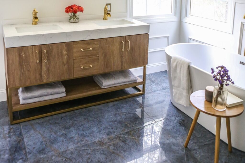 A modern bathroom featuring a wooden vanity with two sinks and gold faucets, a freestanding white bathtub with a towel draped over the side, and a small wooden table holding a vase with purple flowers. The floor is covered in polished blue marble tiles.