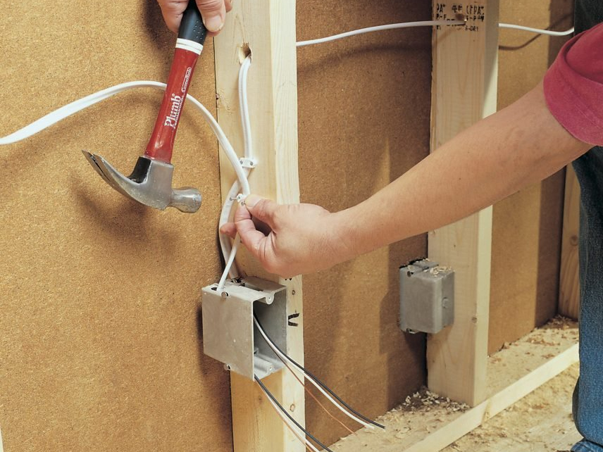 A person is using a hammer to install a staple over electrical wiring on a wooden stud. The wires lead to an electrical box mounted on the stud. The scene appears to be part of a residential construction or remodeling project.