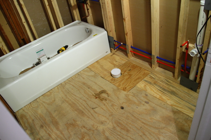 A bathroom under construction with exposed wooden framing and subfloor. A white bathtub installed on the left side, surrounded by loose tools and materials. Pipes are visible on the wall to the right. The floor is unfinished plywood with a circular plumbing fixture.