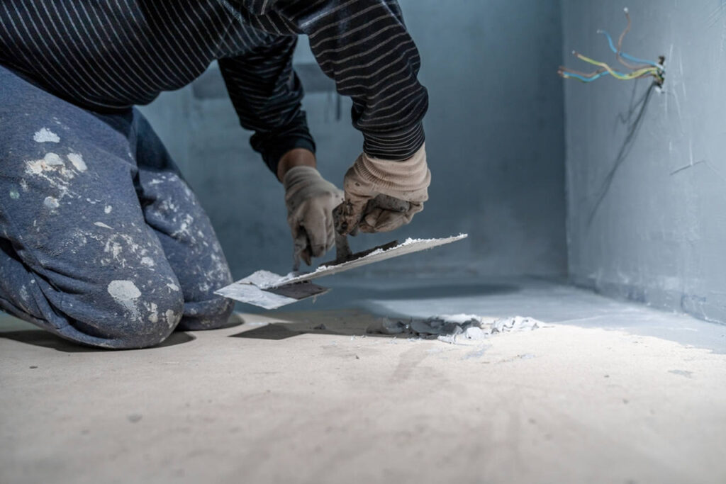 A person, dressed in paint-splattered clothes and wearing gloves, is kneeling on the floor while using a trowel to apply mortar or plaster to a surface. Nearby, exposed electrical wires extend from a wall. The scene suggests ongoing home renovation or construction work.