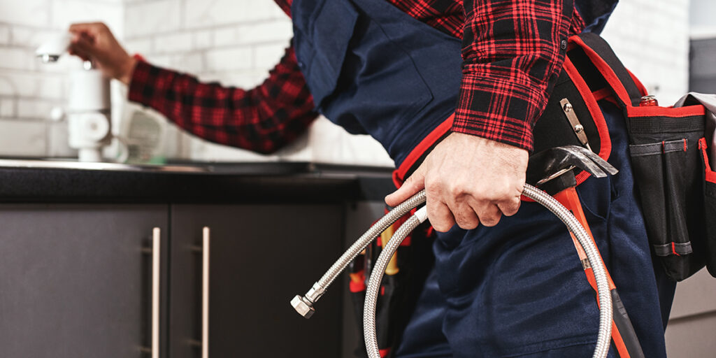 A plumber in a blue uniform and red checkered shirt holds a flexible metal hose and stands in a kitchen. He has a tool belt around his waist with various tools. His other hand is near a sink faucet, suggesting he is about to perform plumbing work.