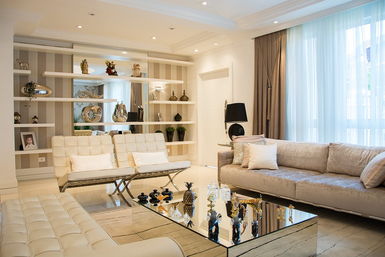 A modern living room with a large beige sofa, two white leather chairs, and a glass coffee table adorned with decorative items. Behind the chairs, there is a wall unit with shelves displaying vases and ornaments. Natural light comes through large windows with sheer curtains.