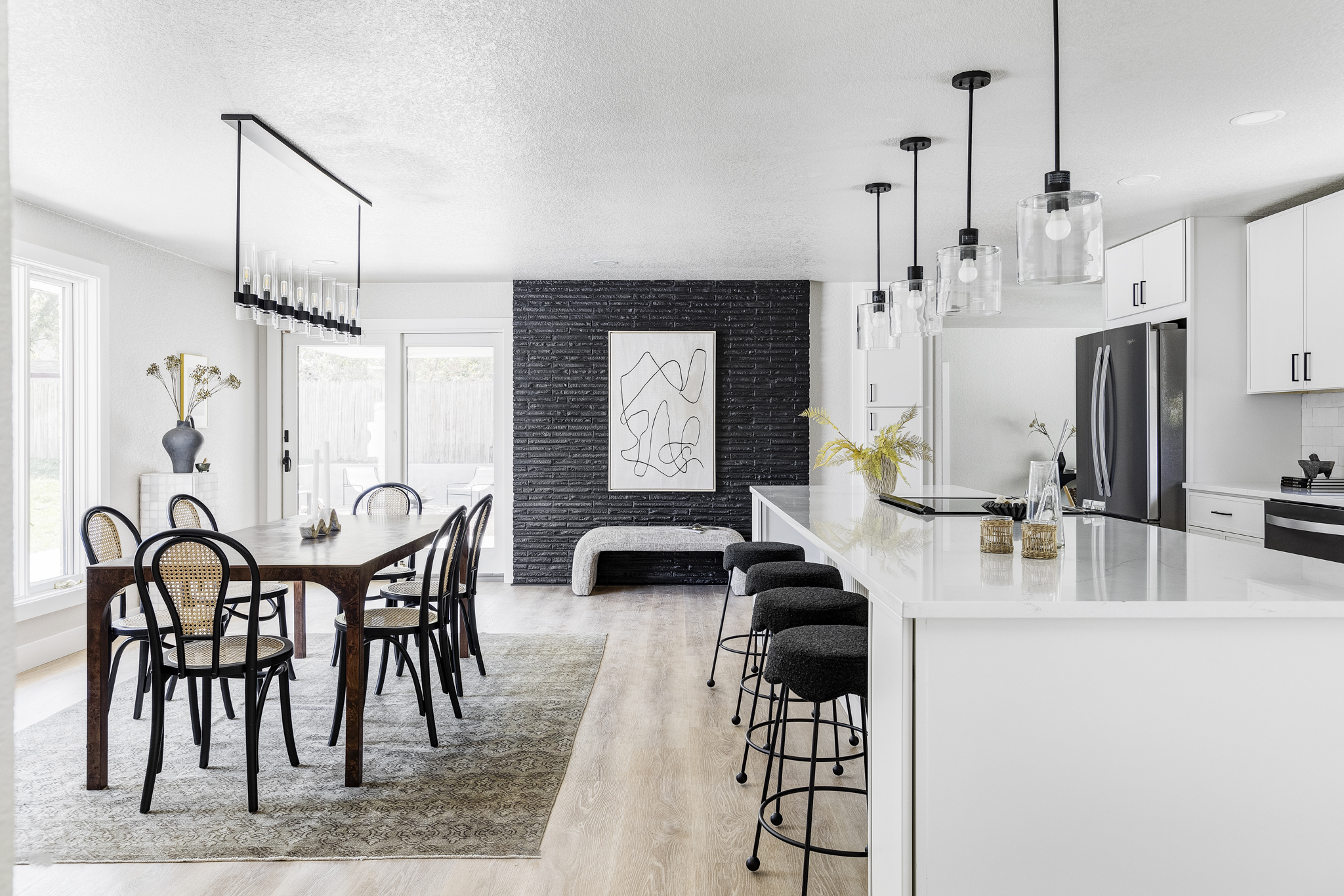 A modern kitchen and dining area with a large white island, black bar stools, and white cabinets. Pendant lights hang above the island. The dining table has black chairs and a vase on top. A black textured wall features abstract art. Bright natural light fills the space.